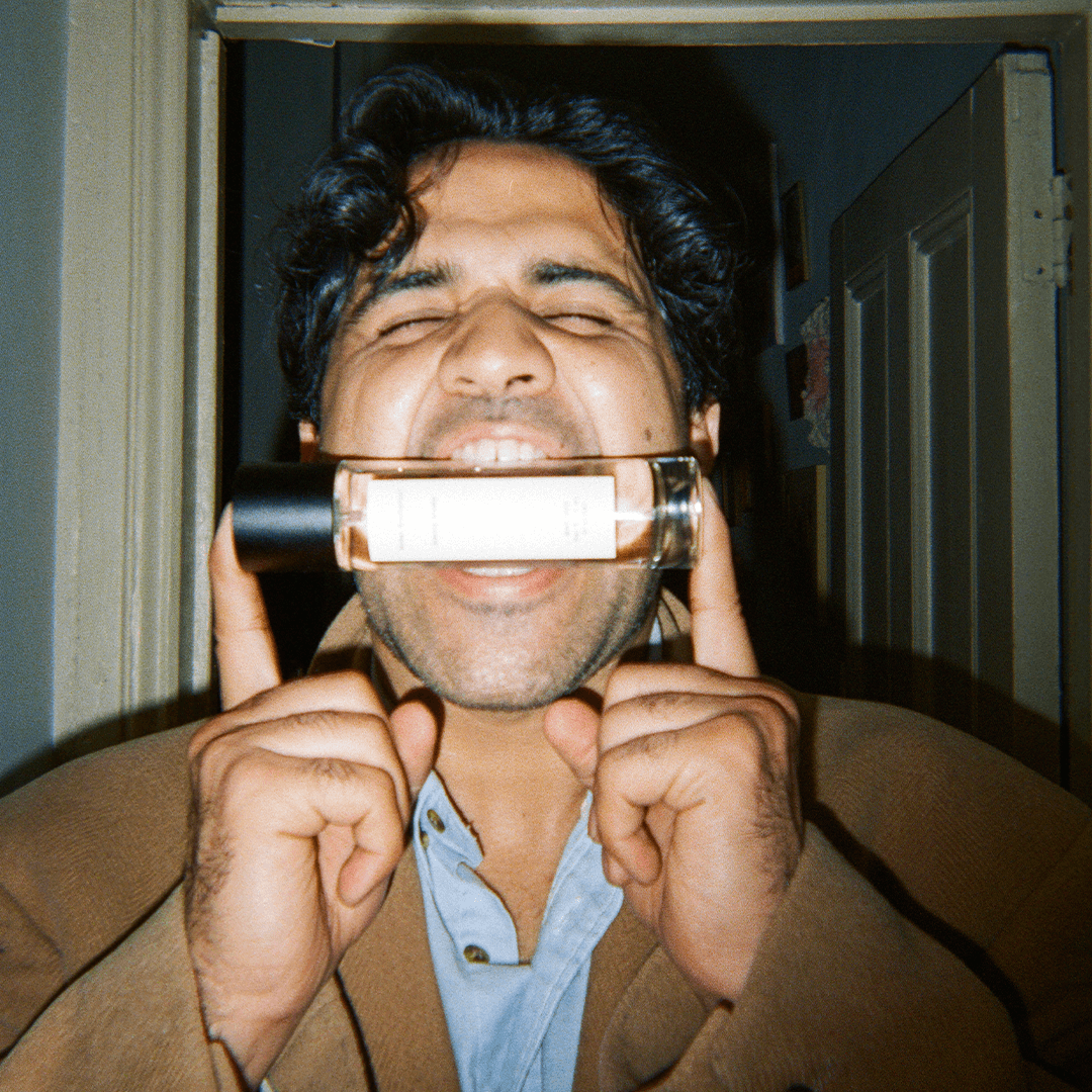 Smiling man holding a perfume bottle between his teeth, showing excitement in a home setting.