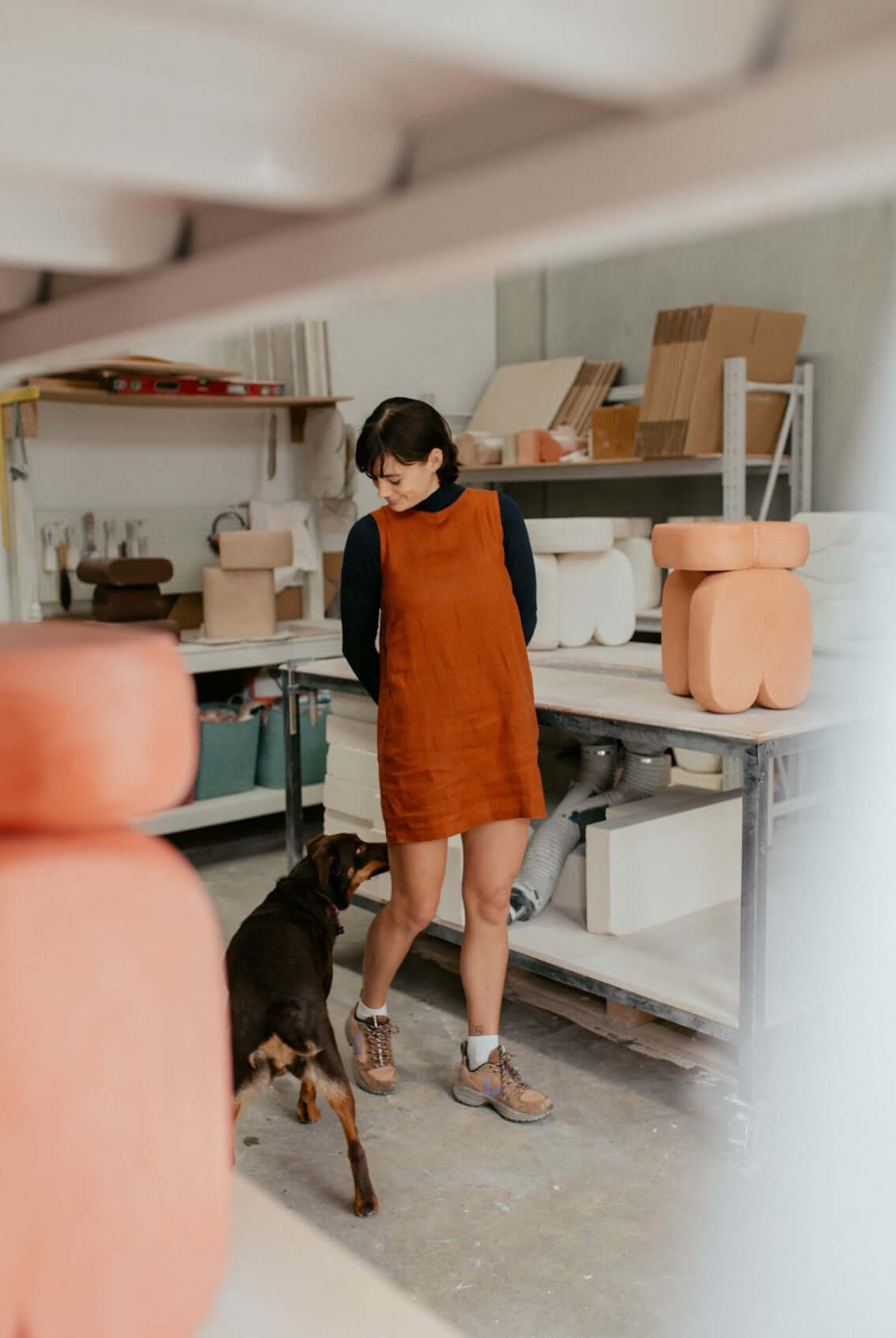 A woman in an orange dress stands in a pottery studio with a dog, surrounded by clay sculptures and tools.
