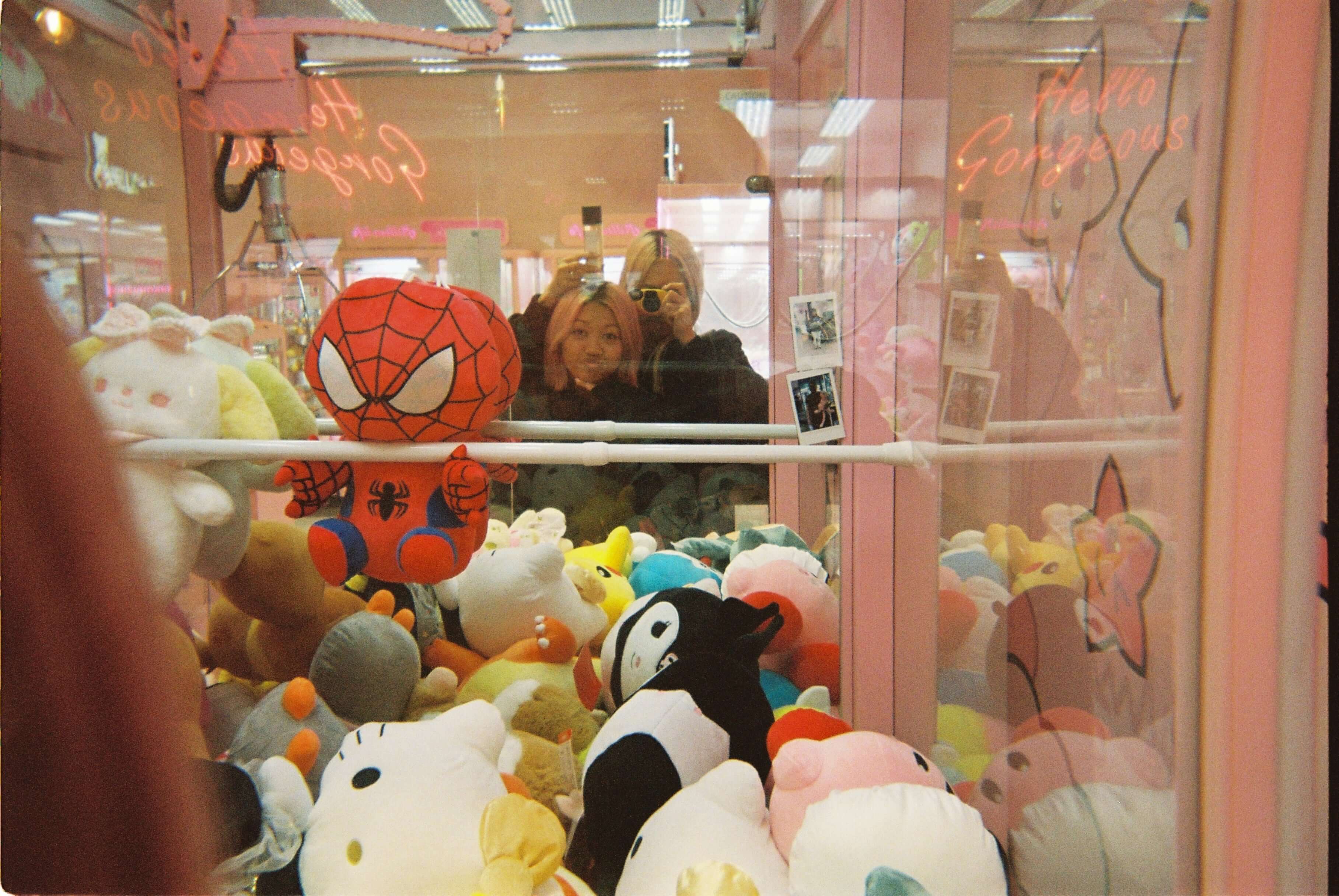 Two friends smiling in front of a claw machine filled with colorful plush toys, including a Spiderman doll.