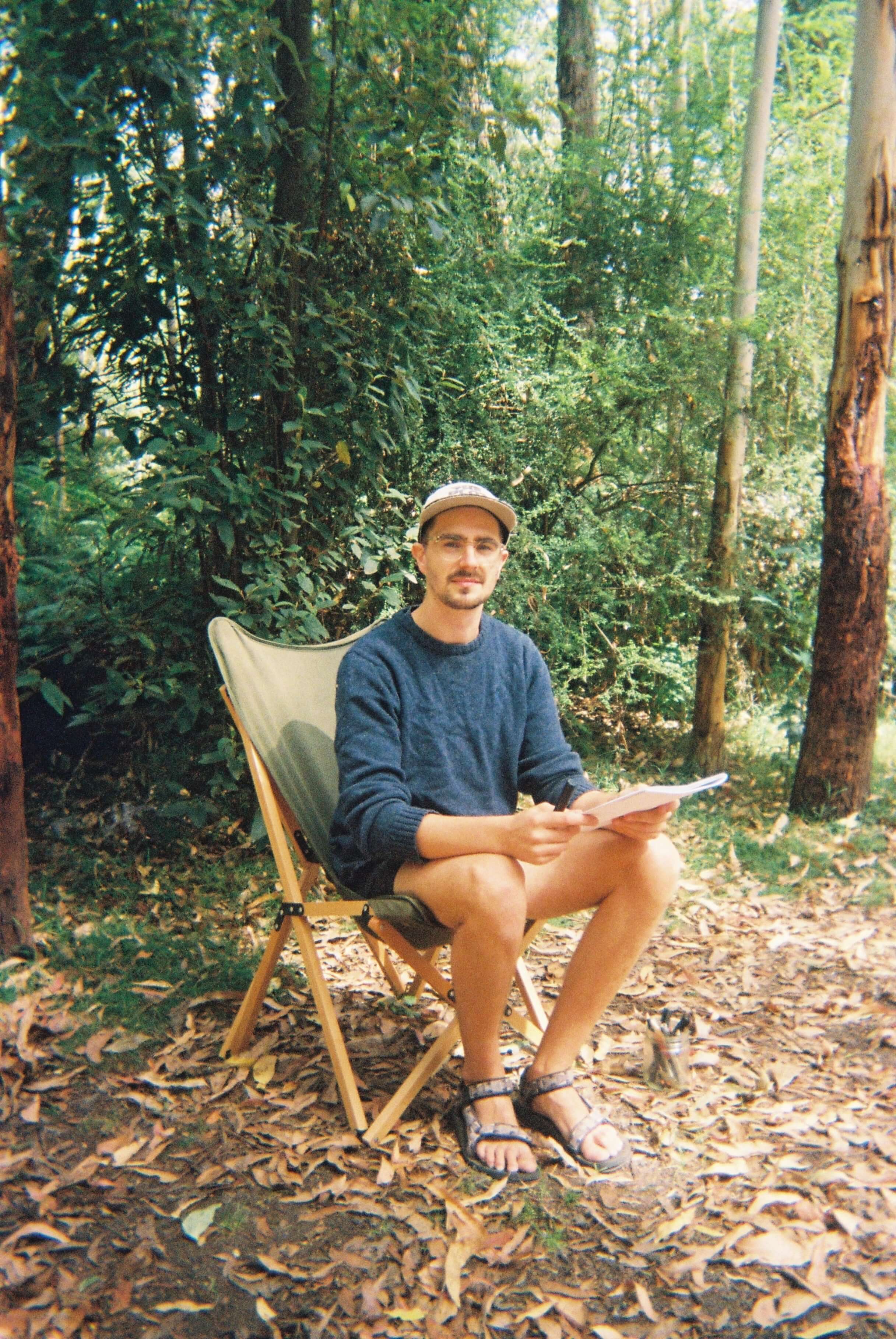 Man sitting on a folding chair in a forest, wearing a sweater and hat, holding a notebook.