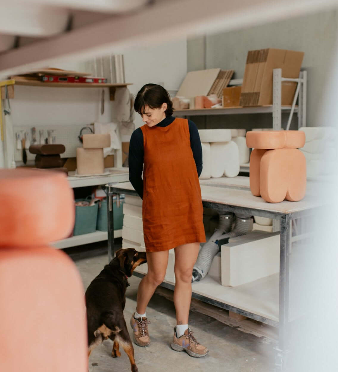 A woman in an orange dress stands in a pottery studio with a dog, surrounded by clay sculptures and tools.