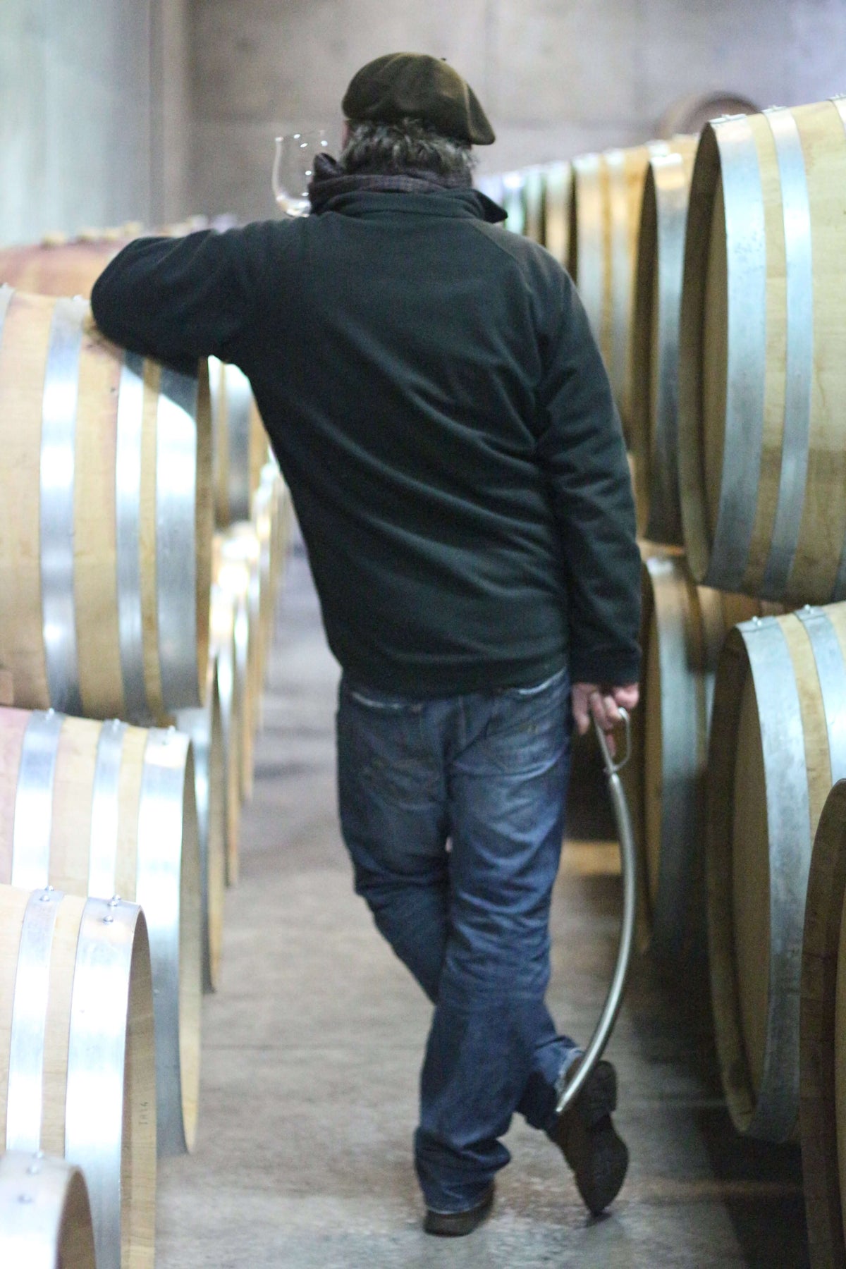Man holding a glass in a winery, leaning against a barrel surrounded by oak casks.