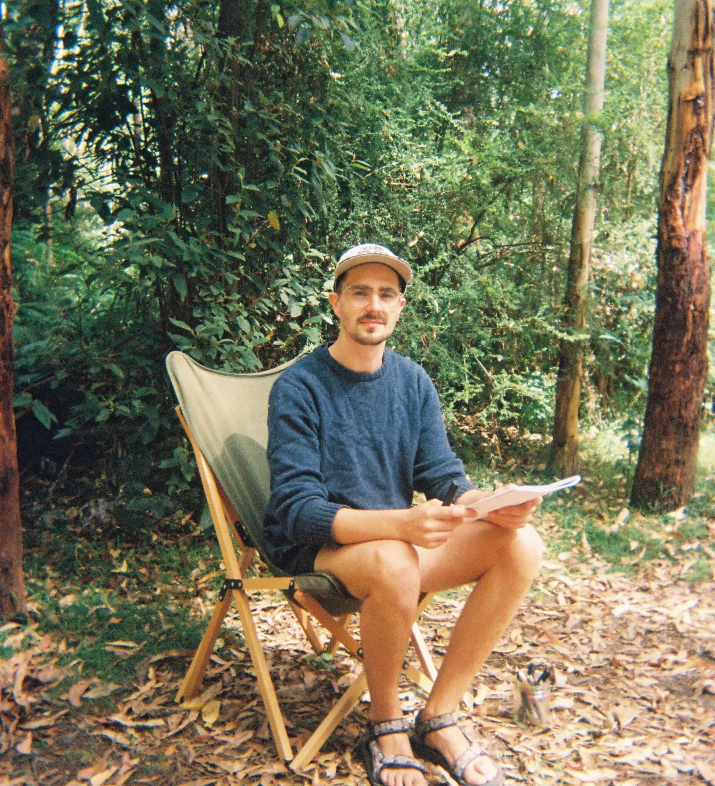 Man sitting on a folding chair in a forest, wearing a sweater and hat, holding a notebook.