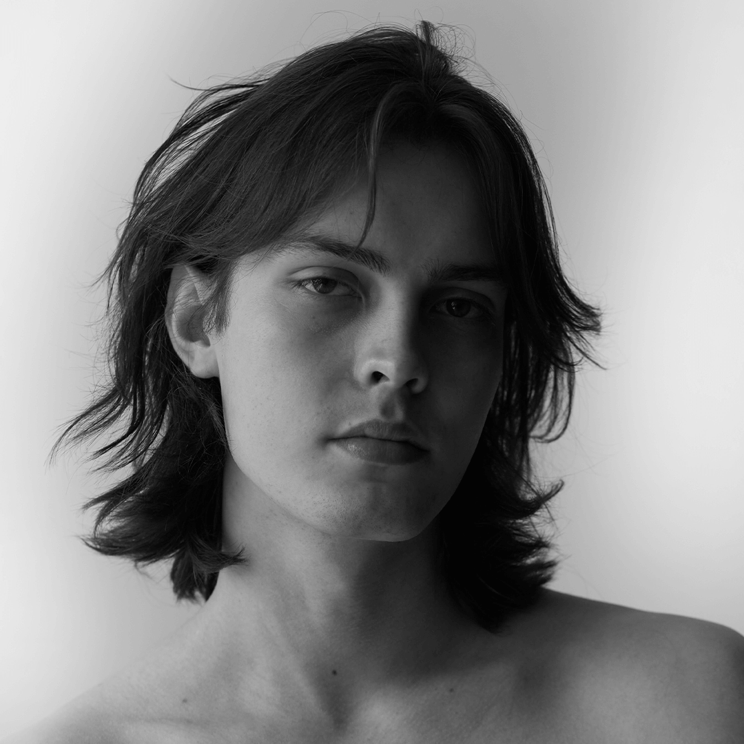 Black and white portrait of a young man with long hair, looking contemplative and direct at the camera.