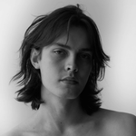 Black and white portrait of a young man with long hair, looking contemplative and direct at the camera.