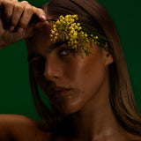 Model holding wattle blossom to her brow against a deep green backdrop, Mihan Aromatics Series Three campaign