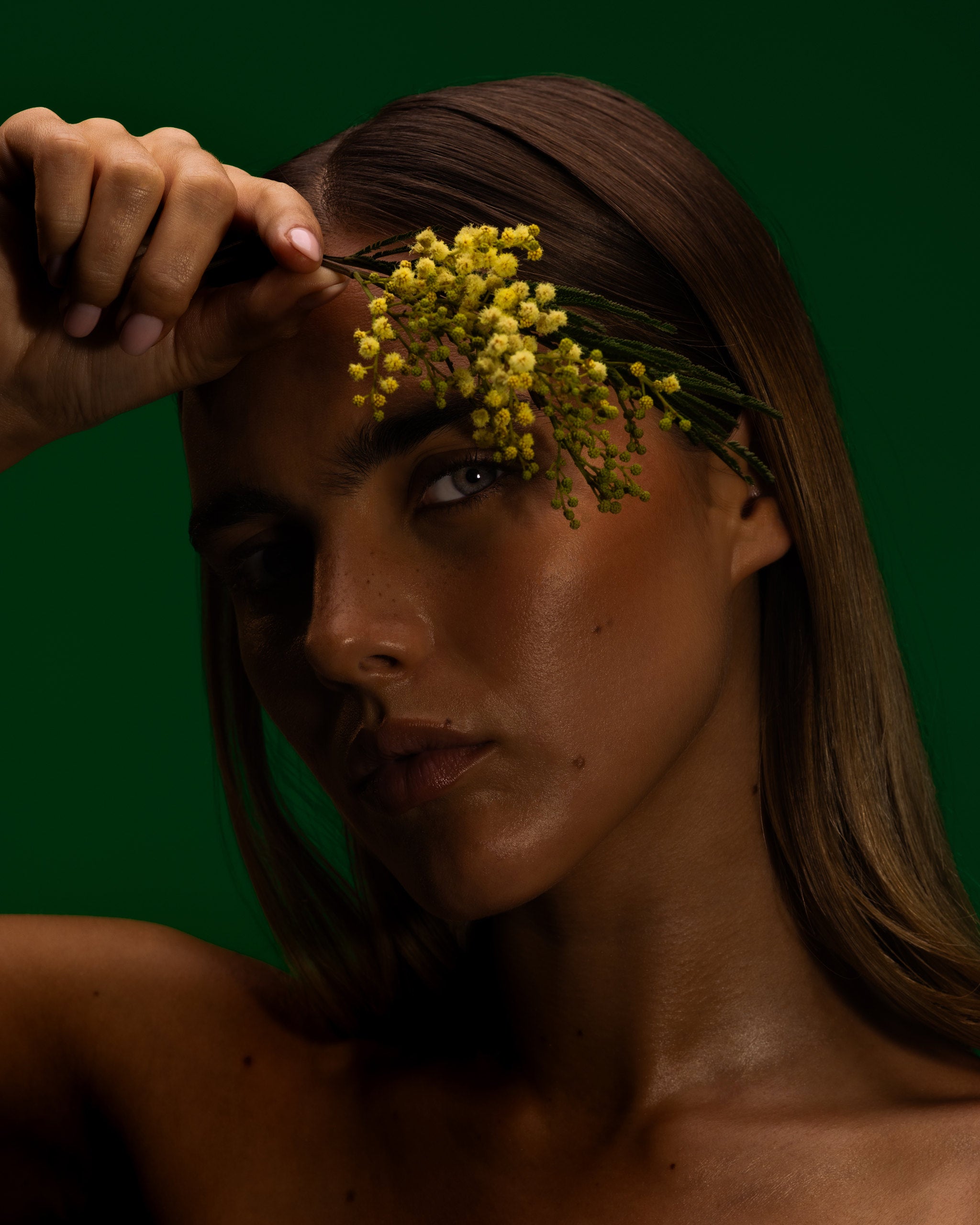 Model holding wattle blossom to her brow against a deep green backdrop, Mihan Aromatics Series Three campaign