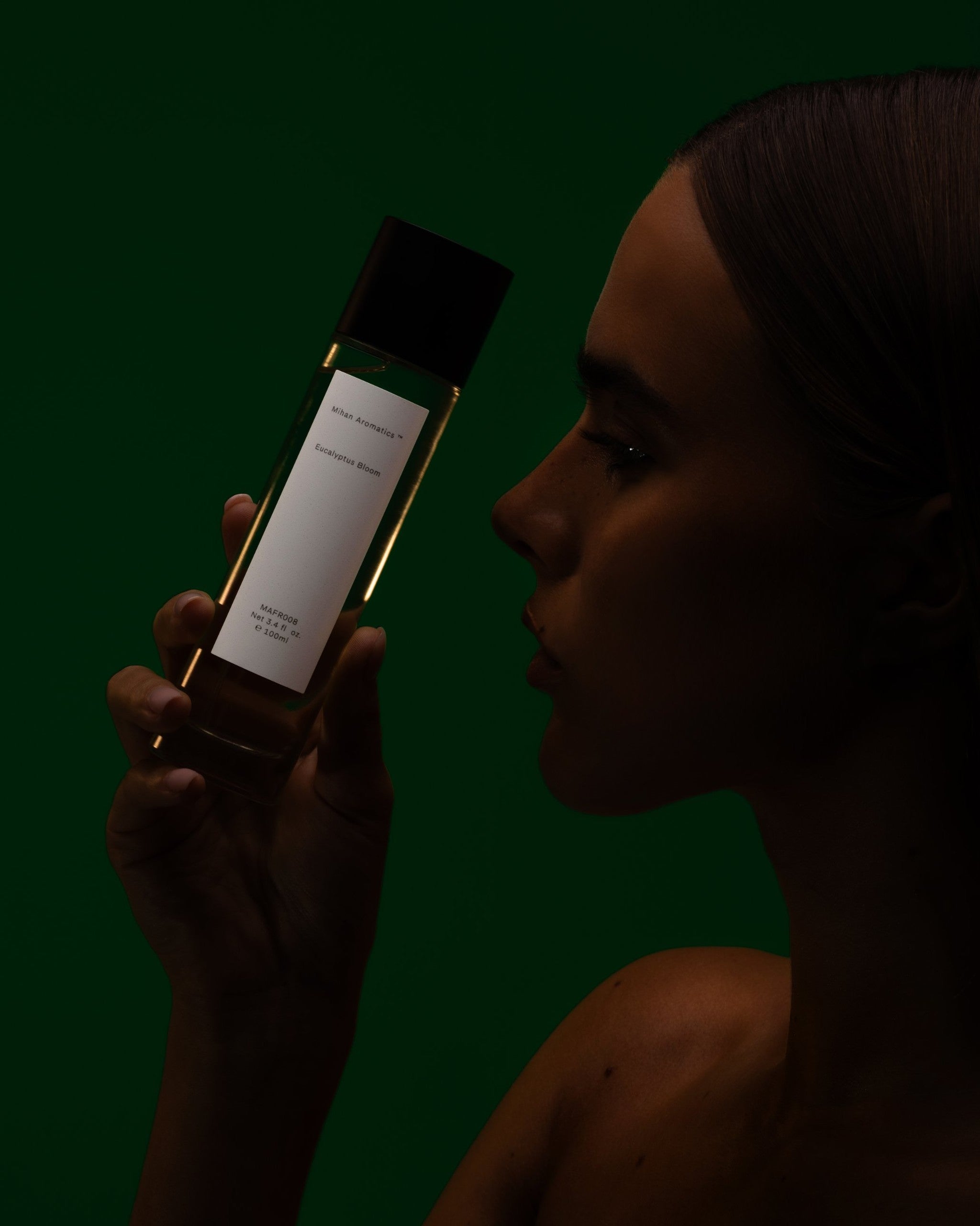 Model in shadow raising a Mihan Aromatics Eucalyptus Bloom parfum bottle to her face against deep green