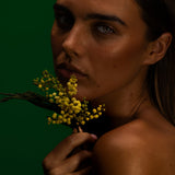 Model holding a spray of wattle to her chin against a deep green backdrop, Mihan Aromatics Eucalyptus Bloom parfum campaign