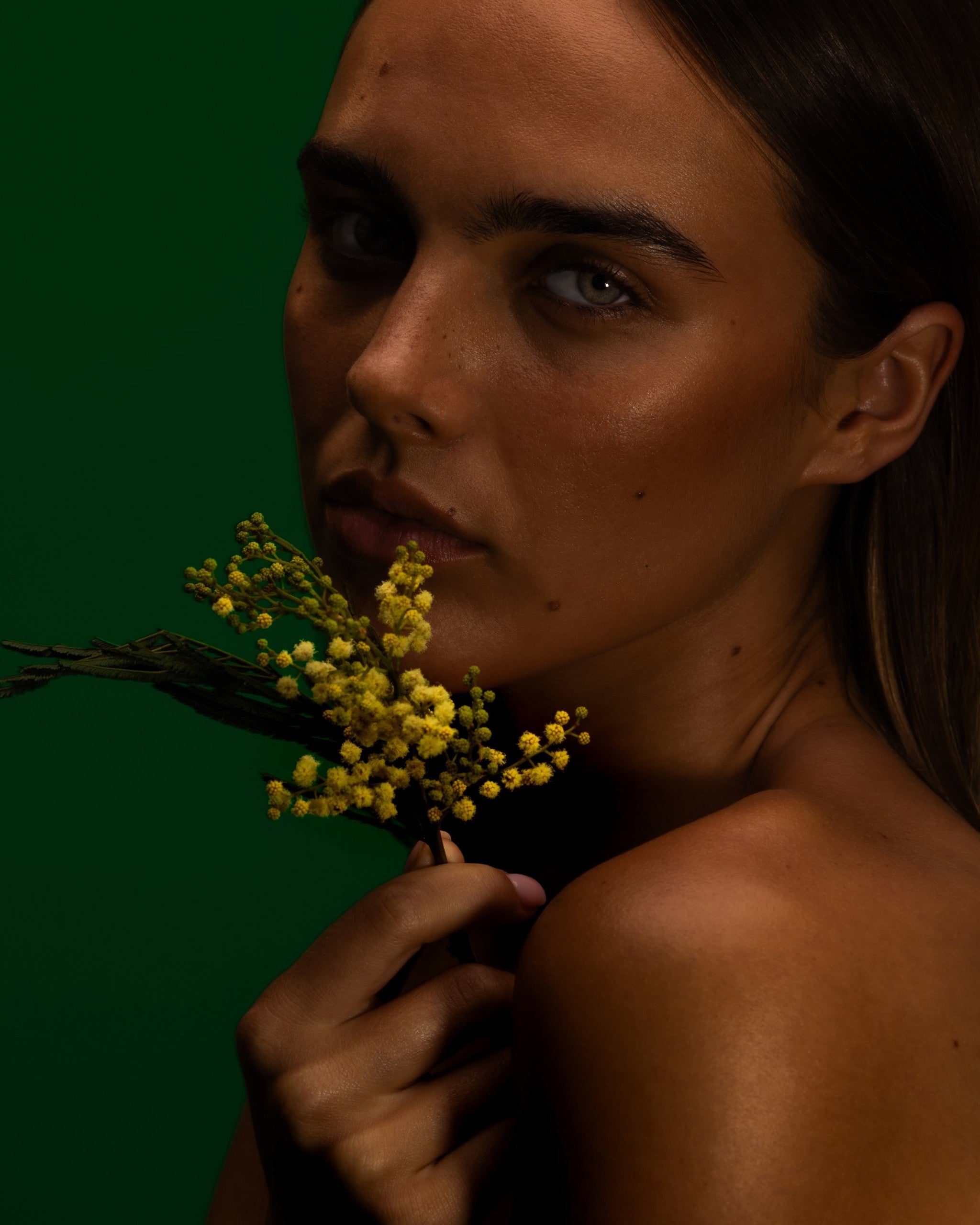 Model holding a spray of wattle to her chin against a deep green backdrop, Mihan Aromatics Eucalyptus Bloom parfum campaign