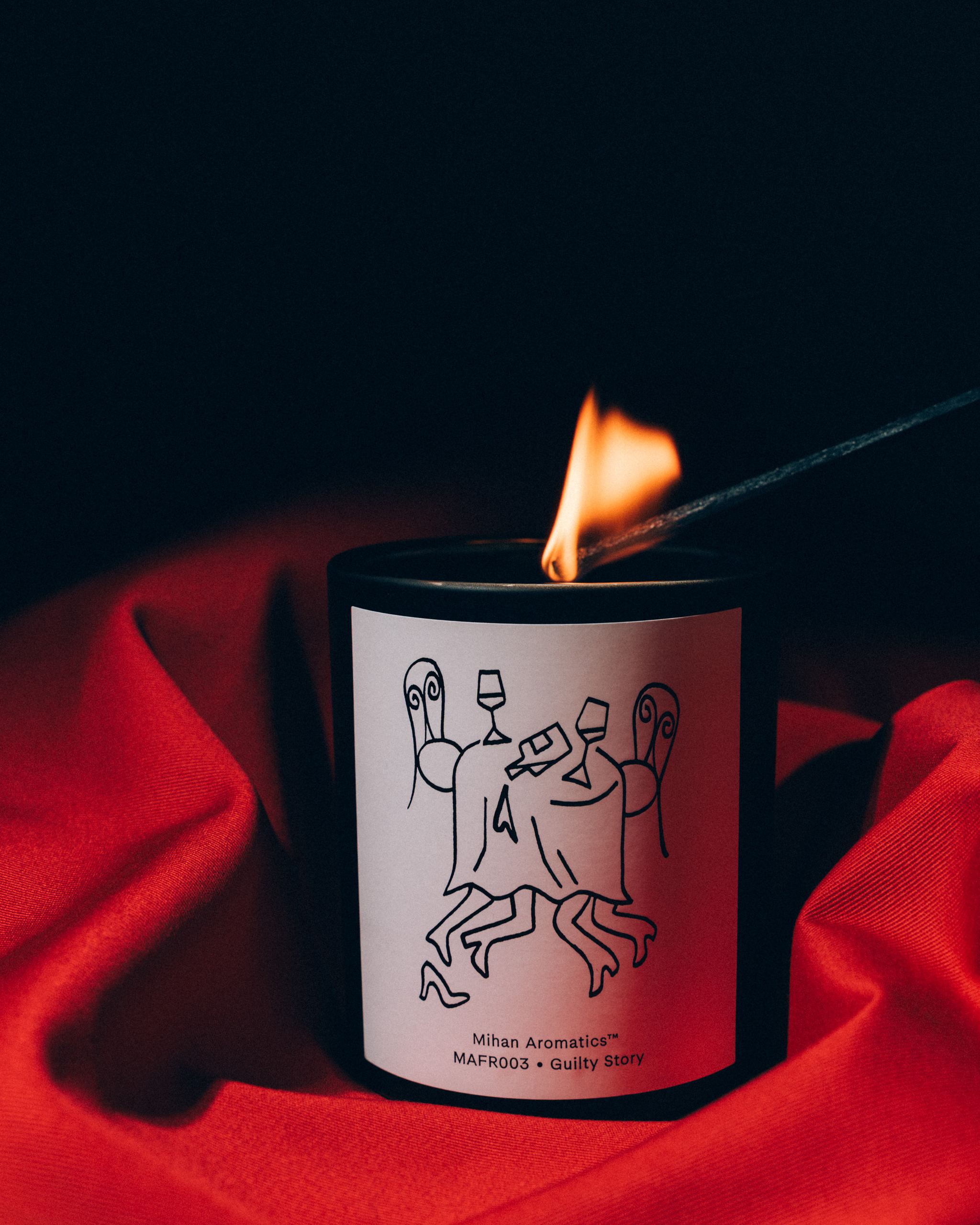Mihan Aromatics Guilty Story candle being lit with a long match, nestled in red fabric