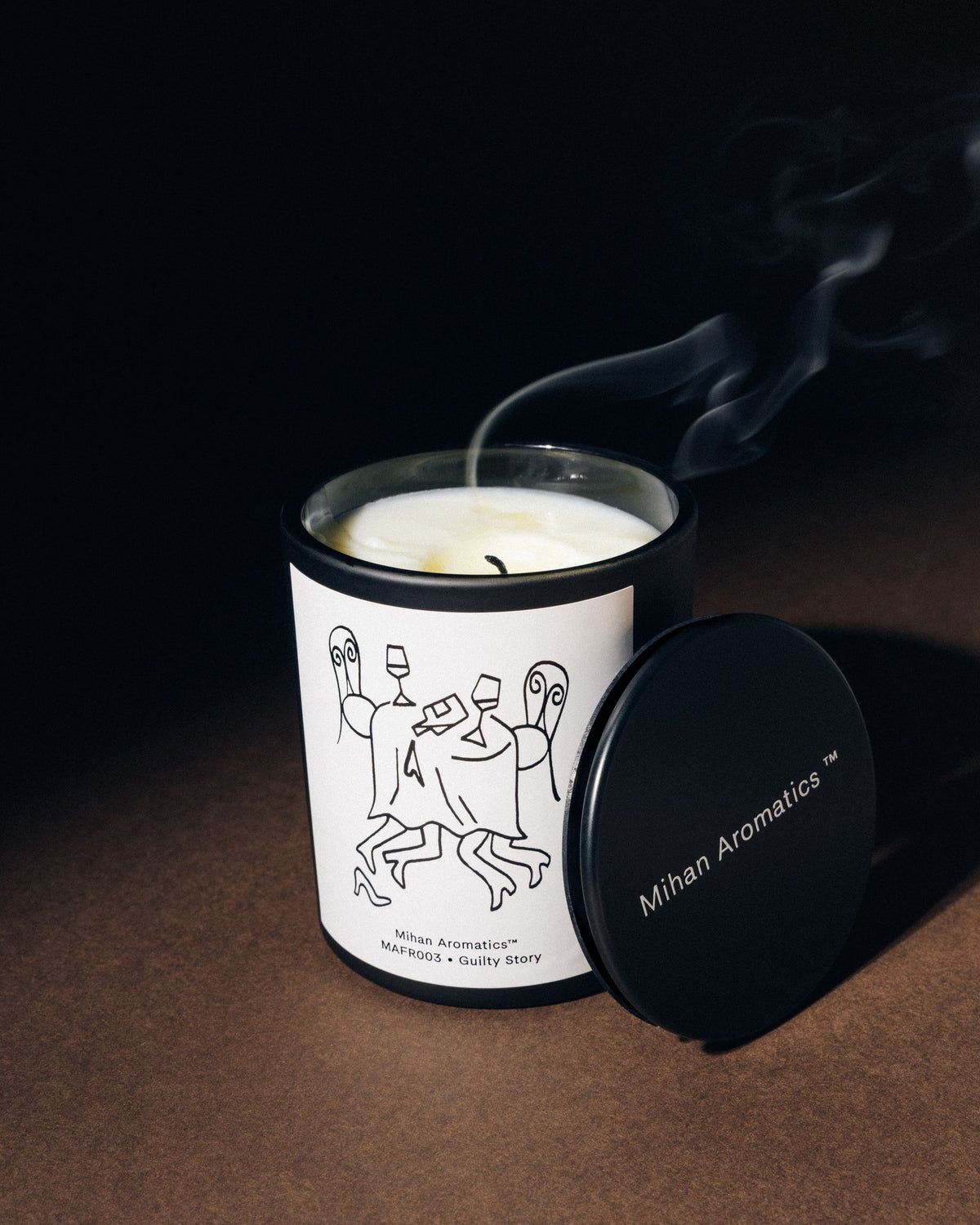 Mihan Aromatics Guilty Story candle with its lid set aside and a trail of smoke rising from the wick