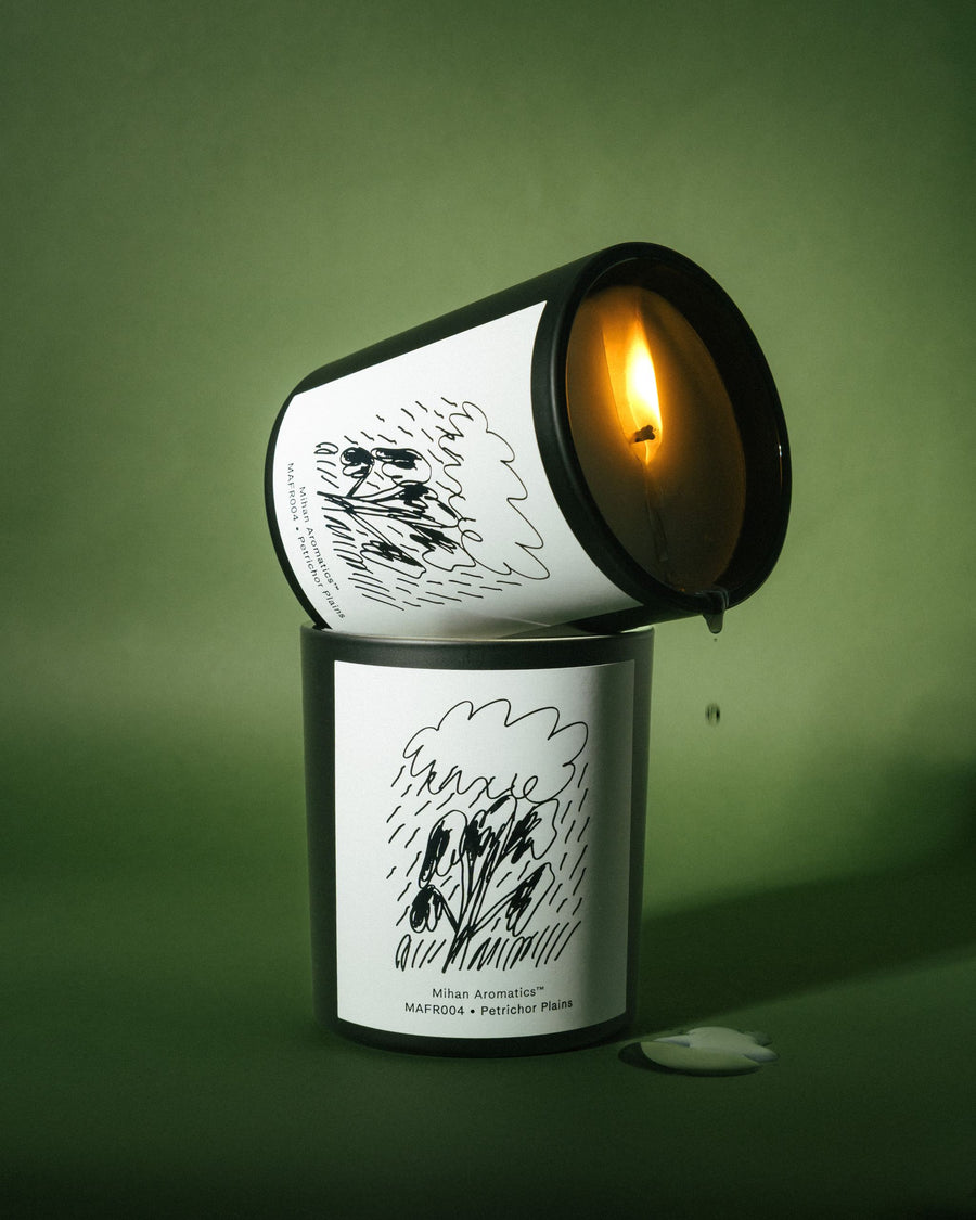 Two stacked Mihan Aromatics Petrichor Plains candles, the upper one lit and tilted with wax dripping