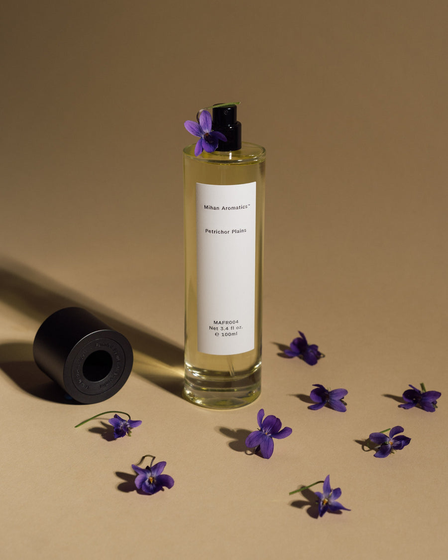 Mihan Aromatics Petrichor Plains parfum 100ml bottle uncapped among scattered violets on a warm sand backdrop