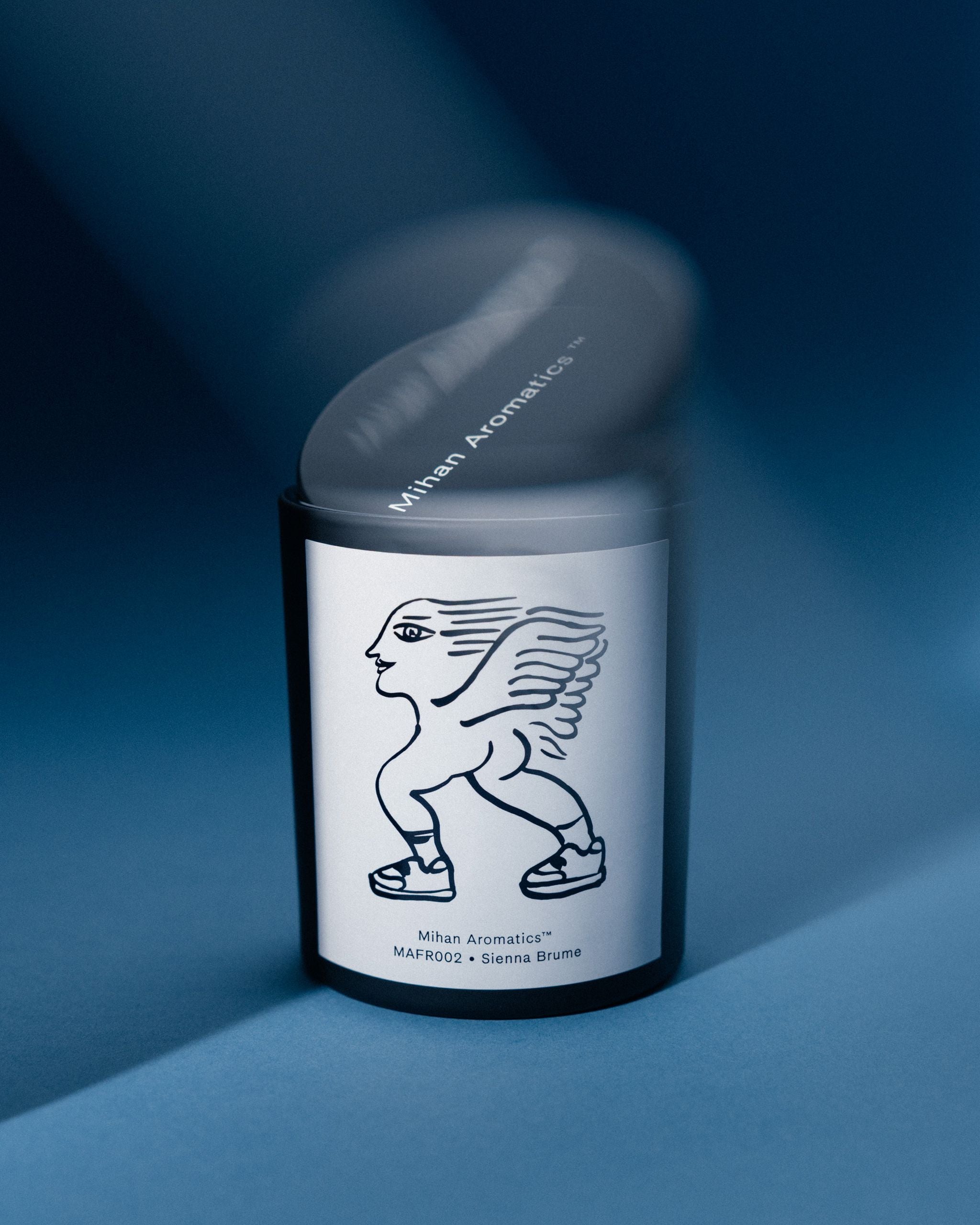 Mihan Aromatics Sienna Brume candle with its lid lifting away in motion on a blue backdrop