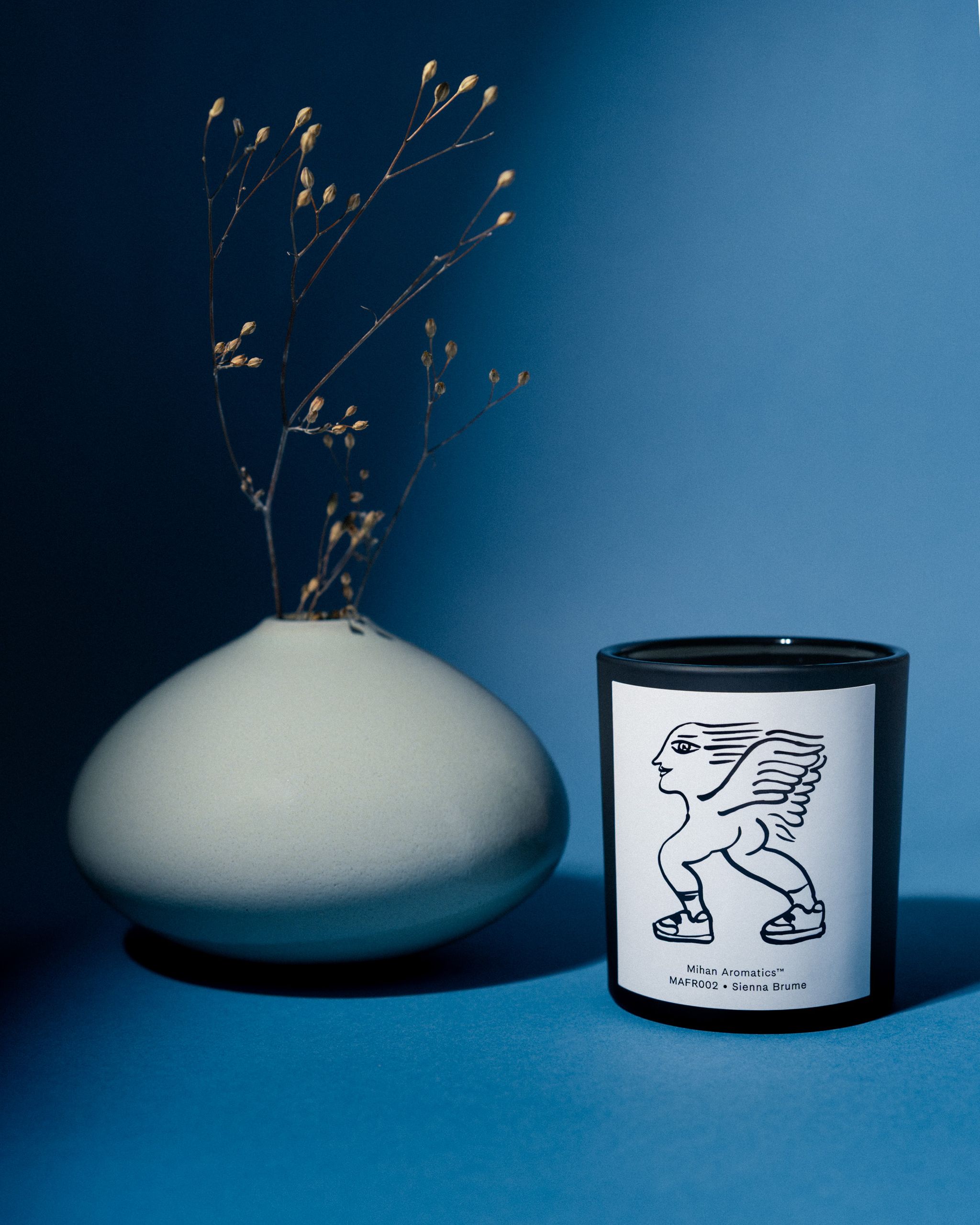 Mihan Aromatics Sienna Brume candle beside a ceramic vase of dried stems on a blue backdrop